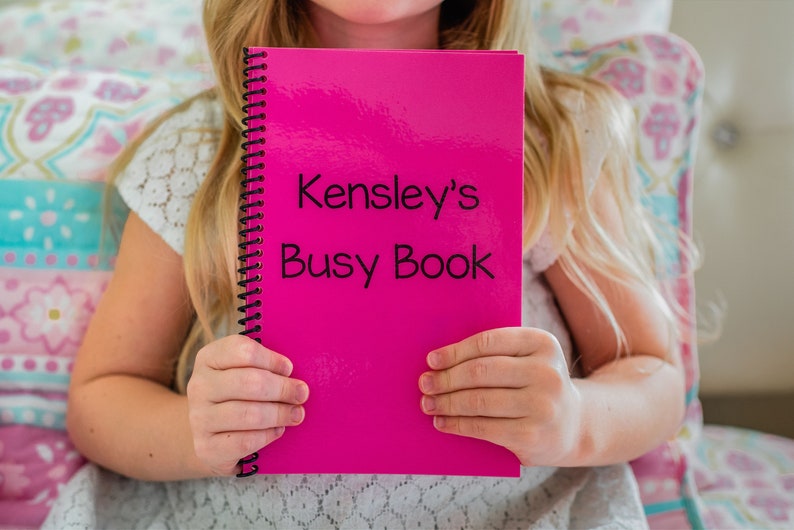 young girl holding her pink personalized laminated busy book. The book says Kensleys busy book.