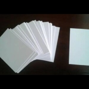 May include: A stack of white rectangular cards fanned out, with a single card to the right. The cards are plain and appear to be made of paper or cardstock. The background is a dark, solid color.