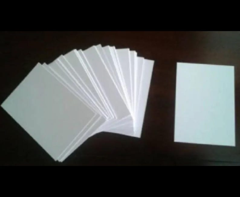 May include: A stack of white rectangular cards fanned out, with a single card to the right. The cards are plain and appear to be made of paper or cardstock. The background is a dark, solid color.