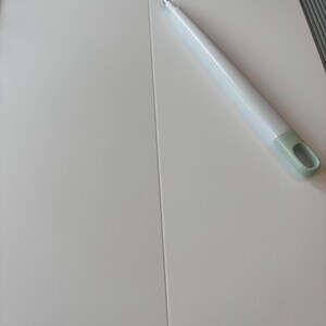 May include: A white paper trimmer with a light green handle rests on a white surface. The trimmer has a gray cutting guide and a sharp blade. The surface is divided into two sections by a vertical crease.