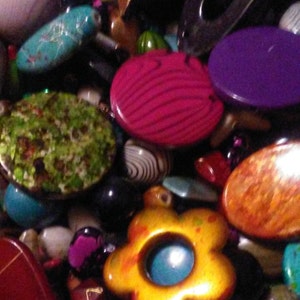 May include: A close-up of a collection of beads and decorative items. The image features a variety of shapes and colours, including turquoise, pink, purple, and yellow. Some beads have patterns, such as stripes and floral designs, creating a diverse and visually appealing mix.