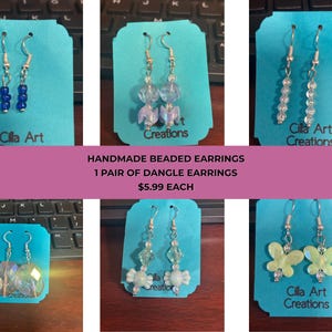 May include: A collection of handmade beaded dangle earrings displayed on teal cards. The earrings feature various designs, including blue beads, clear beads, and butterfly shapes. The text "Cilla Art Creations" is visible on some cards.