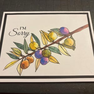 May include: A watercolor illustration of an olive branch with green leaves and colorful olives in shades of yellow, purple, and orange. The words "I'M Sorry" are written in elegant script above the branch, set against a white background with a black border.