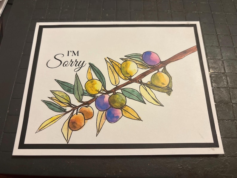 May include: A watercolor illustration of an olive branch with green leaves and colorful olives in shades of yellow, purple, and orange. The words "I'M Sorry" are written in elegant script above the branch, set against a white background with a black border.