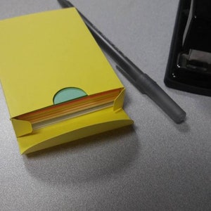 May include: A yellow paper holder with a stack of colorful papers peeking out. A gray pen and a black stapler are also on the gray surface. Office supplies.