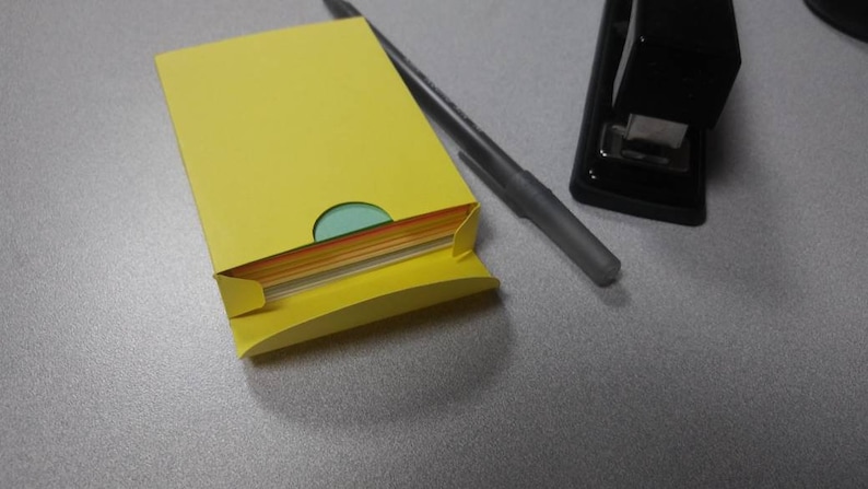 May include: A yellow paper holder with a stack of colorful papers peeking out. A gray pen and a black stapler are also on the gray surface. Office supplies.