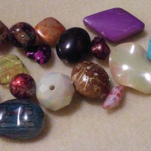 May include: A selection of colourful beads in various shapes and sizes. The beads include a mix of colours such as purple, teal, brown, orange, and black. Some beads are faceted, while others are smooth and rounded. These beads are suitable for jewellery making.