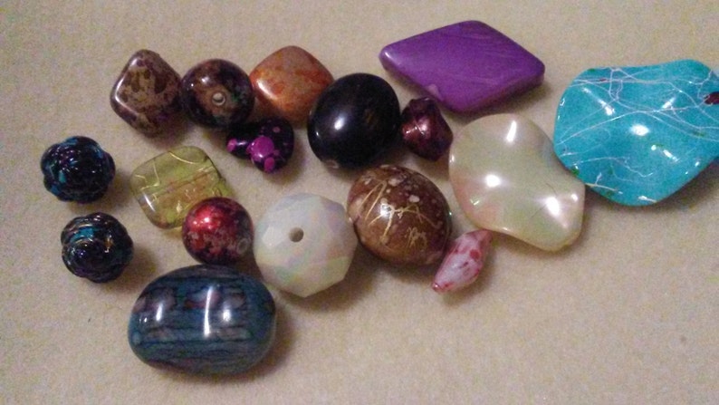 May include: A selection of colourful beads in various shapes and sizes. The beads include a mix of colours such as purple, teal, brown, orange, and black. Some beads are faceted, while others are smooth and rounded. These beads are suitable for jewellery making.