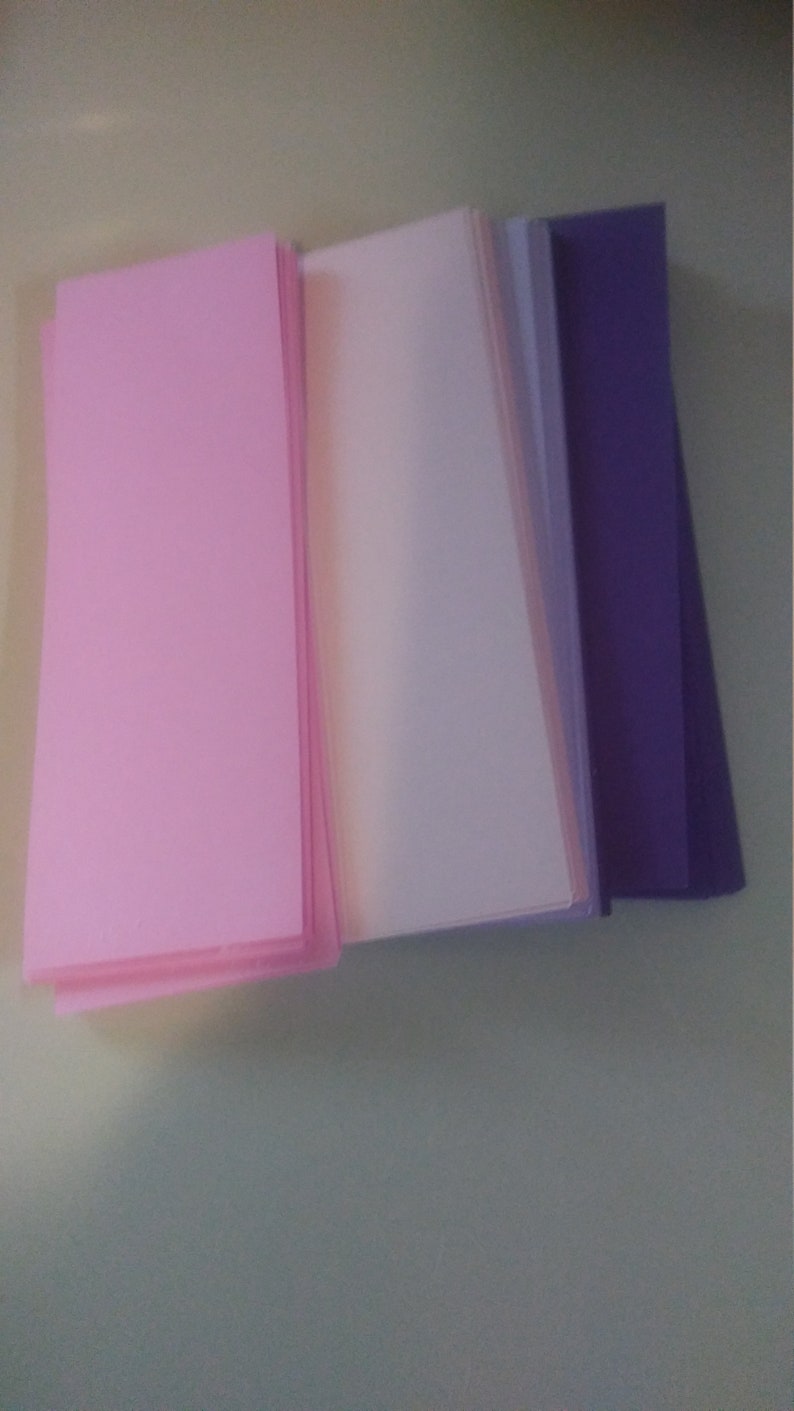 K&ouml;nnte beinhalten: Ein Stapel rechteckiger Papierb&ouml;gen in den Farbt&ouml;nen Rosa, Off-White und Lila. Die Papiere sind ordentlich angeordnet, mit den rosa B&ouml;gen links, gefolgt von Off-White- und lila B&ouml;gen rechts. Das Papier scheint glatt und von mittlerer Dicke zu sein.