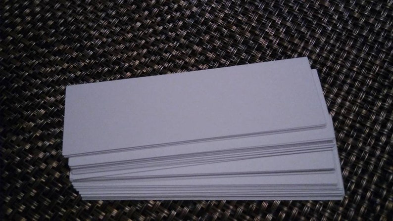 K&ouml;nnte beinhalten: Ein Stapel rechteckiger, hellgrauer Blankokarten. Die Karten sind ordentlich gestapelt, die Kanten ausgerichtet. Der Hintergrund ist eine dunkle, strukturierte Oberfl&auml;che. Die Karten scheinen aus Papier oder Karton zu sein.