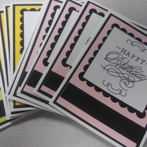 May include: A stack of birthday cards with a pink and black design. The cards have a scalloped border and the words "Happy Birthday" in elegant script. Some cards have handwritten notes.