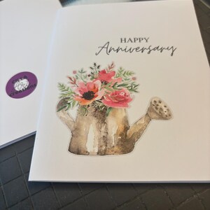 May include: A white greeting card with the words "HAPPY Anniversary" in black script. The card features a watercolor illustration of a watering can filled with pink and red flowers and green foliage. A second card is visible in the background.