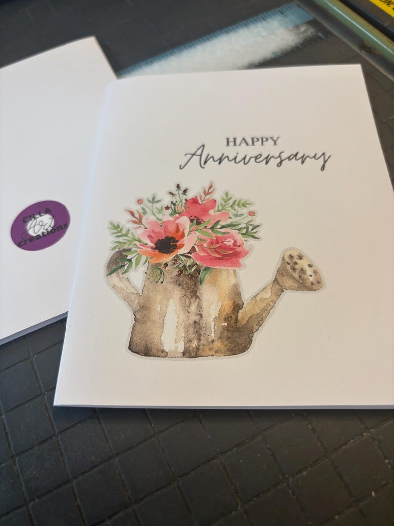 May include: A white greeting card with the words "HAPPY Anniversary" in black script. The card features a watercolor illustration of a watering can filled with pink and red flowers and green foliage. A second card is visible in the background.