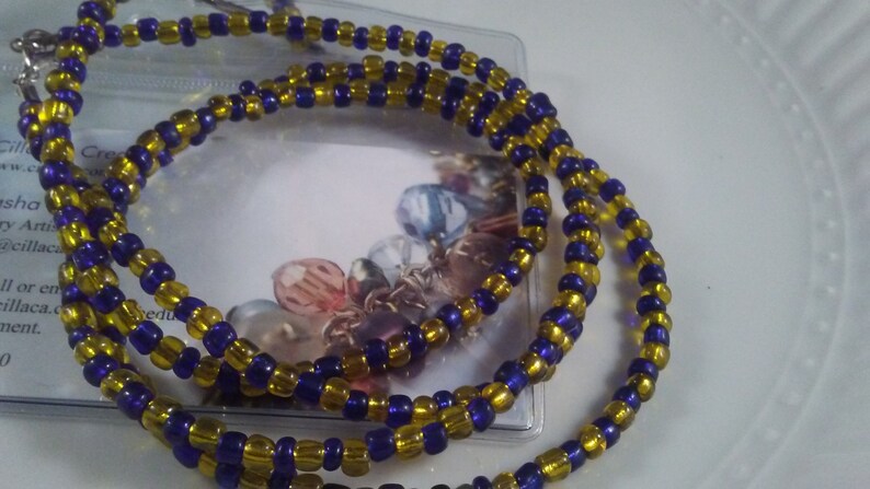 May include: A beaded necklace featuring alternating gold and dark blue beads. The necklace is made of small, round beads strung together, creating a layered effect. The necklace is displayed on a white surface.