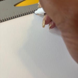 May include: A white marker is used to draw a line on a white piece of paper. A gray and yellow ruler is used as a guide. The image is a close-up of the process of drawing a line.