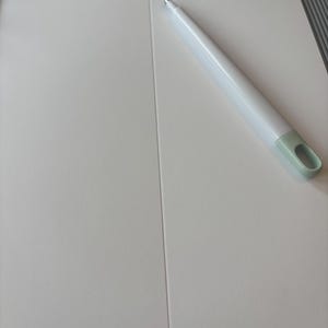 May include: A white paper trimmer with a light green handle and a white pen. The trimmer is on a white surface with a scoring guide. The pen is white with a silver tip.