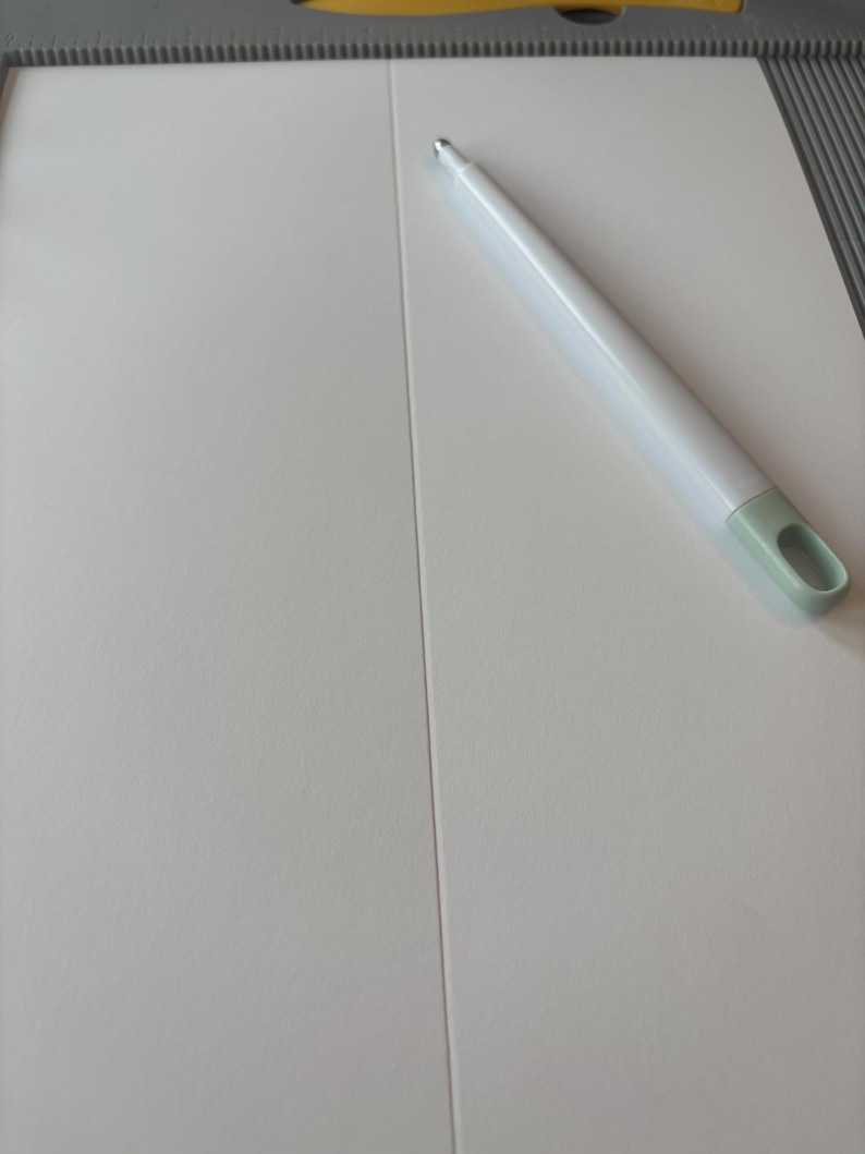 May include: A white paper trimmer with a light green handle and a white pen. The trimmer is on a white surface with a scoring guide. The pen is white with a silver tip.