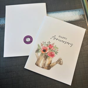 May include: A white greeting card with the words "Happy Anniversary" in script. The card features a watercolor illustration of a watering can overflowing with pink and red flowers. The back of the card has a purple logo.