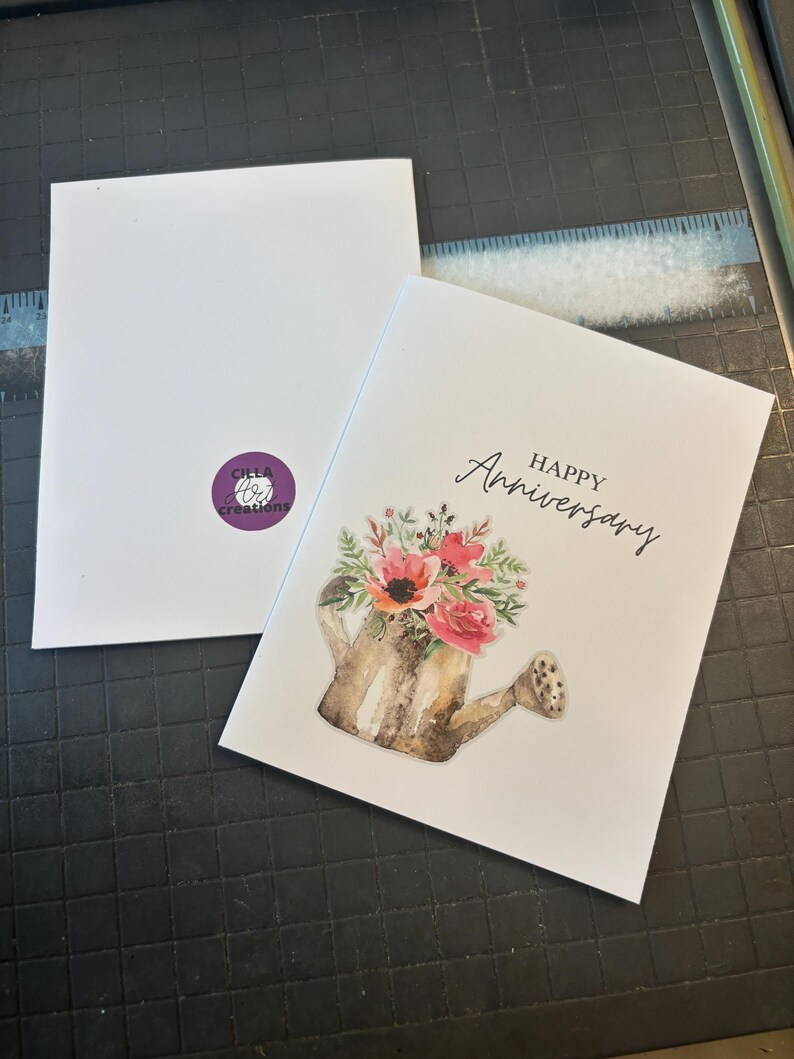 May include: A white greeting card with the words "Happy Anniversary" in script. The card features a watercolor illustration of a watering can overflowing with pink and red flowers. The back of the card has a purple logo.