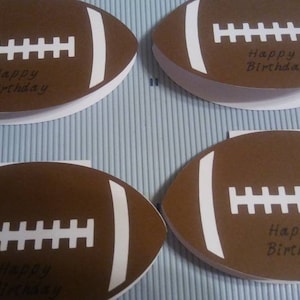 Peut inclure: Quatre cartes de v&oelig;ux en forme de ballon de football marron avec des d&eacute;tails blancs et le texte "Happy Birthday". Les cartes sont dispos&eacute;es sur une surface ray&eacute;e bleu clair et blanc, pour un anniversaire sportif.