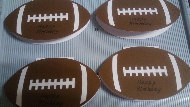 Peut inclure: Quatre cartes de v&oelig;ux en forme de ballon de football marron avec des d&eacute;tails blancs et le texte "Happy Birthday". Les cartes sont dispos&eacute;es sur une surface ray&eacute;e bleu clair et blanc, pour un anniversaire sportif.