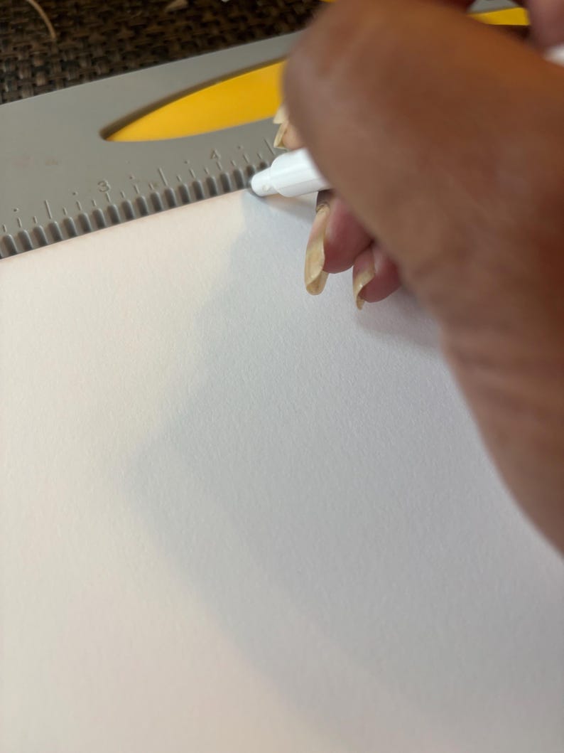 May include: A white marker is used to draw a line on a white piece of paper. A gray and yellow ruler is positioned at the top of the paper, with measurements in inches. The image is a close-up shot.