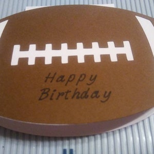 Peut inclure: Une carte de v&oelig;ux en forme de ballon de football marron avec des rayures et des coutures blanches. Les mots "Happy Birthday" sont &eacute;crits en script noir au centre. La carte est sur une surface bleu clair.