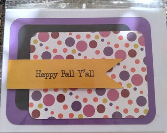 May include: A greeting card with a white background, featuring layers of purple, black, and a patterned paper with colorful polka dots. A yellow banner across the front reads "Happy Fall Y'all". The card is sealed in a clear plastic sleeve.