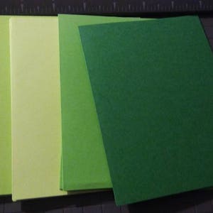 May include: A collection of rectangular green and yellow paper sheets arranged in a fan shape. The shades range from light yellow-green to a deep forest green. The paper is on a dark surface with a cutting guide.