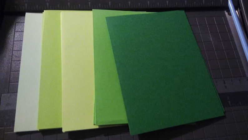 May include: A collection of rectangular green and yellow paper sheets arranged in a fan shape. The shades range from light yellow-green to a deep forest green. The paper is on a dark surface with a cutting guide.