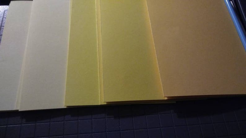 May include: A stack of rectangular sheets of paper in varying shades of yellow and tan. The colors range from a pale cream to a deeper, golden hue. The papers are neatly arranged, suggesting a stationery or craft supply.