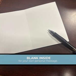 May include: An open, blank greeting card with a black pen resting on the right side. The card is white, and the pen is black with a silver tip. The text at the bottom reads "BLANK INSIDE for your own personal message."