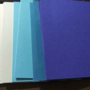 May include: A stack of rectangular paper sheets in shades of white, light blue, and dark blue. The papers are arranged in a vertical row, showcasing a gradient of colors. The papers are on a dark surface.