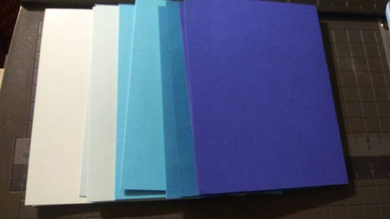 May include: A stack of rectangular paper sheets in shades of white, light blue, and dark blue. The papers are arranged in a vertical row, showcasing a gradient of colors. The papers are on a dark surface.