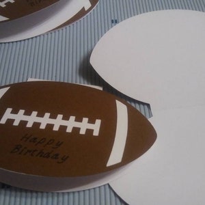 Peut inclure: Cartes de v&oelig;ux en forme de ballon de football marron et blanc avec l'inscription "Happy Birthday". Les cartes sont ouvertes et ferm&eacute;es, avec un int&eacute;rieur blanc. Les cartes sont sur une surface ray&eacute;e bleu clair et blanc.