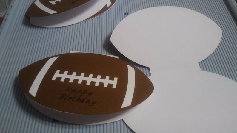 Peut inclure: Cartes de v&oelig;ux en forme de ballon de football marron et blanc avec l'inscription "Happy Birthday". Les cartes sont ouvertes et ferm&eacute;es, avec un int&eacute;rieur blanc. Les cartes sont sur une surface ray&eacute;e bleu clair et blanc.
