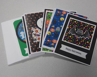 May include: A stack of Halloween-themed greeting cards. The cards feature various designs, including bats, pumpkins, and witches. One card has the text "Happy Halloween" on a banner. The cards are in various colors, including black, orange, and green.