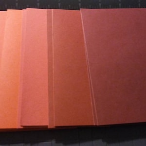 May include: A stack of rectangular paper sheets in varying shades of red and orange. The colors range from light pink to deep red. The sheets are neatly aligned on a cutting mat, with a blue cutting tool visible in the corner.