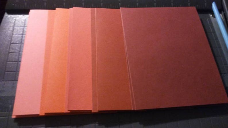 May include: A stack of rectangular paper sheets in varying shades of red and orange. The colors range from light pink to deep red. The sheets are neatly aligned on a cutting mat, with a blue cutting tool visible in the corner.