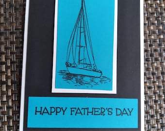 May include: A handmade Father's Day card featuring a teal rectangle with the text "HAPPY FATHER'S DAY". The card has a black background with a teal rectangle in the center, showcasing a black sailboat illustration.