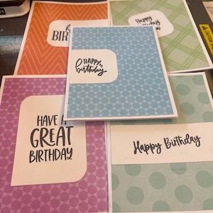 Könnte beinhalten: Eine Sammlung von fünf handgefertigten Grußkarten mit verschiedenen Mustern und Farben. Die Karten zeigen Texte wie Happy Birthday und Have a Great Birthday in schwarzer Tinte auf weißen Etiketten. Die Muster umfassen orangefarbene Zickzacklinien, grünes Karomuster, blaue Dreiecke, violette Sechsecke und hellblaue Punkte.