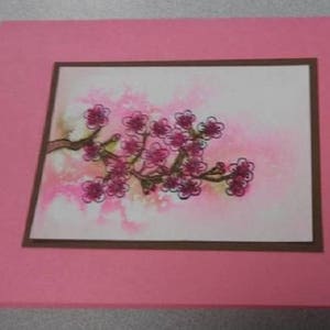 May include: A pink greeting card featuring a watercolor painting of a flowering branch with pink blossoms. The artwork is framed with a brown border and set against a soft pink background.