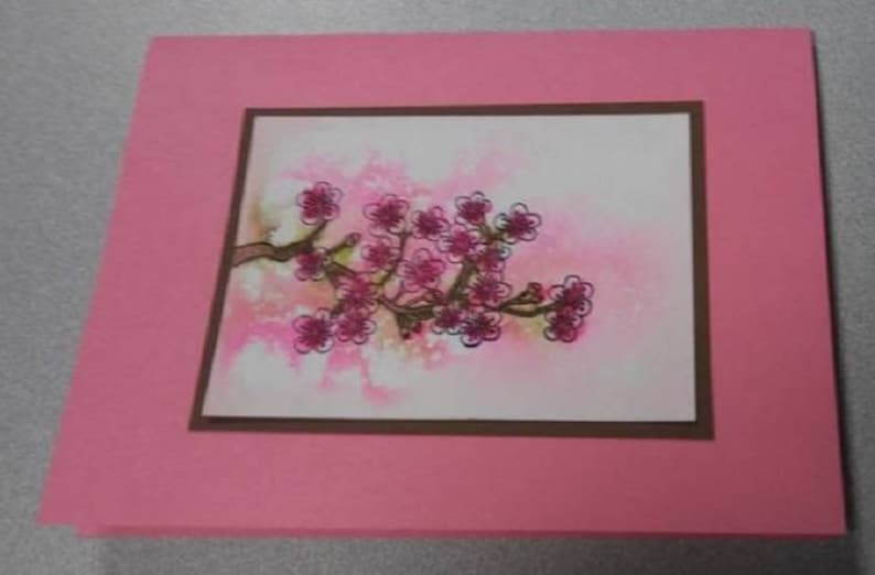 May include: A pink greeting card featuring a watercolor painting of a flowering branch with pink blossoms. The artwork is framed with a brown border and set against a soft pink background.