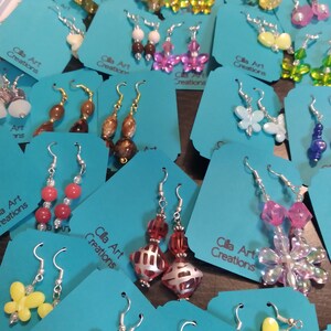 May include: A collection of handmade earrings displayed on teal tags. The earrings feature various designs, including beaded drops, butterfly shapes, and floral patterns. The text "Cilla Art Creations" is visible on the tags. The earrings are in various colors.