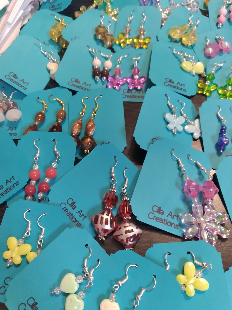 May include: A collection of handmade earrings displayed on teal tags. The earrings feature various designs, including beaded drops, butterfly shapes, and floral patterns. The text "Cilla Art Creations" is visible on the tags. The earrings are in various colors.