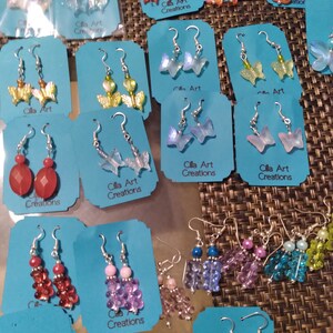 May include: A collection of handmade earrings displayed on blue cards. The earrings feature various designs, including butterfly shapes, beaded clusters, and gemstone drops. The text "Cilla Art Creations" is visible on the cards.