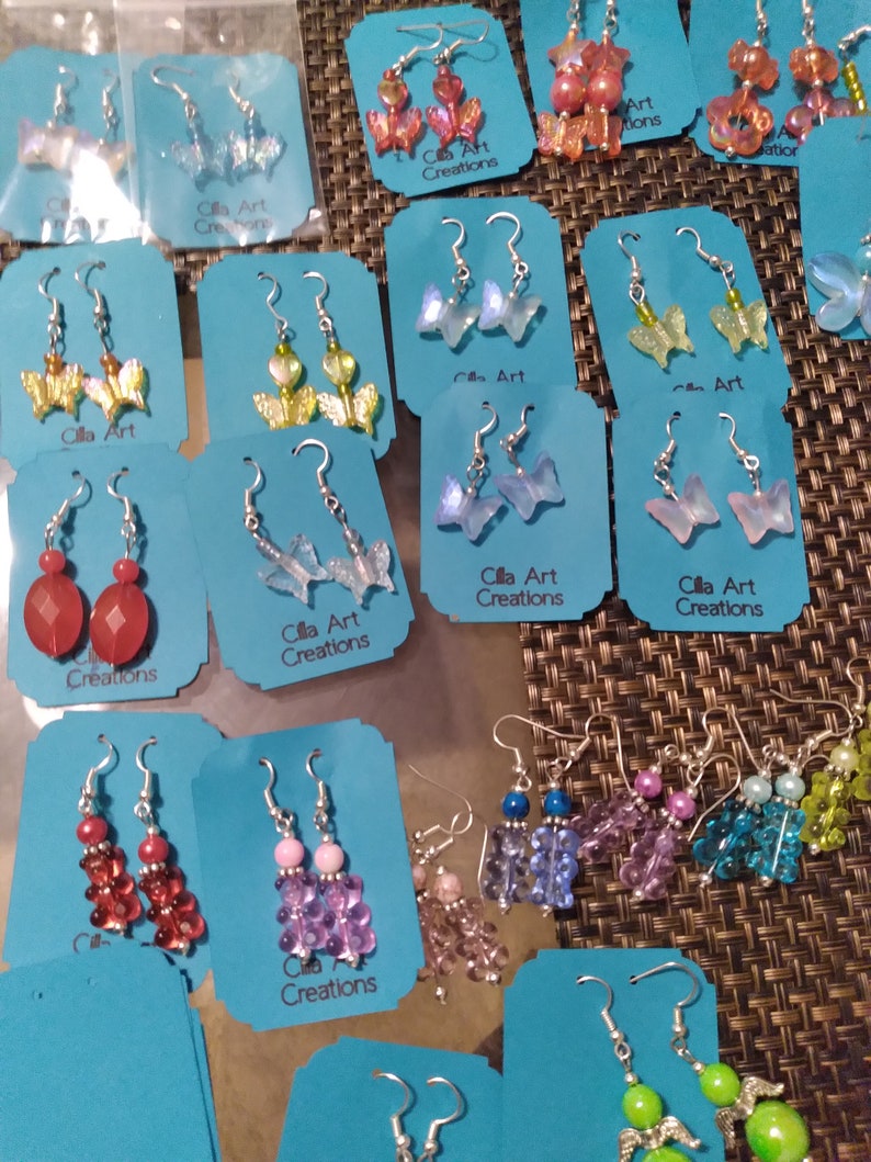 May include: A collection of handmade earrings displayed on blue cards. The earrings feature various designs, including butterfly shapes, beaded clusters, and gemstone drops. The text "Cilla Art Creations" is visible on the cards.
