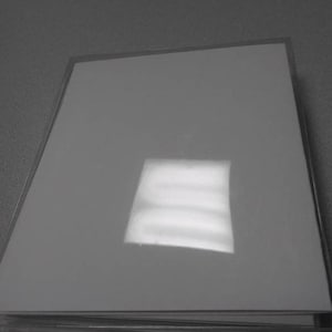 Op de afbeelding: Een stapel witte, glanzende papieren vellen, mogelijk fotopapier of karton, in een doorzichtige plastic hoes. Het papier reflecteert licht, waardoor een helder vierkant ontstaat. De afbeelding is in grijstinten.