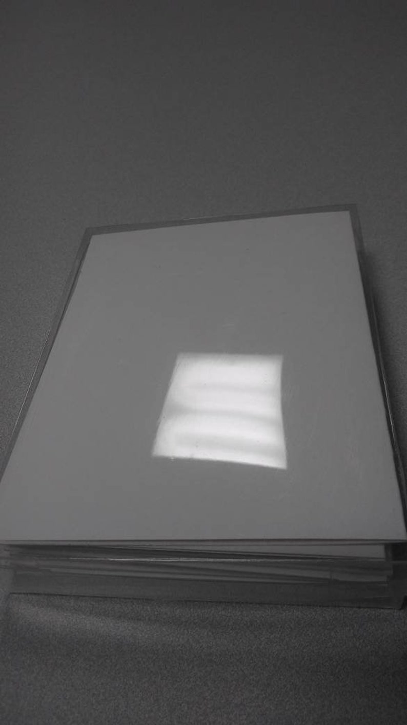 Op de afbeelding: Een stapel witte, glanzende papieren vellen, mogelijk fotopapier of karton, in een doorzichtige plastic hoes. Het papier reflecteert licht, waardoor een helder vierkant ontstaat. De afbeelding is in grijstinten.