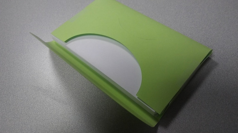 K&ouml;nnte beinhalten: Eine hellgr&uuml;ne CD-H&uuml;lle aus Papier mit einer teilweise sichtbaren wei&szlig;en CD. Die H&uuml;lle ist ge&ouml;ffnet und gibt die Disc frei. Die H&uuml;lle liegt auf einer grauen Oberfl&auml;che.